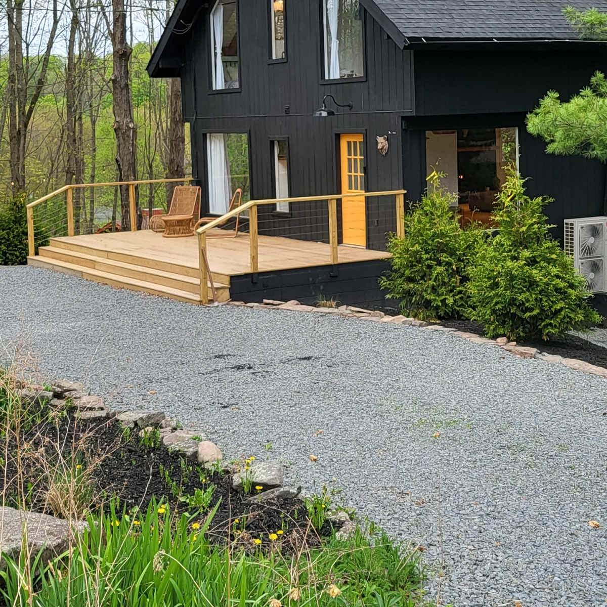 A black wooden house with large windows and a yellow door sits beside a gravel driveway, surrounded by greenery and trees.