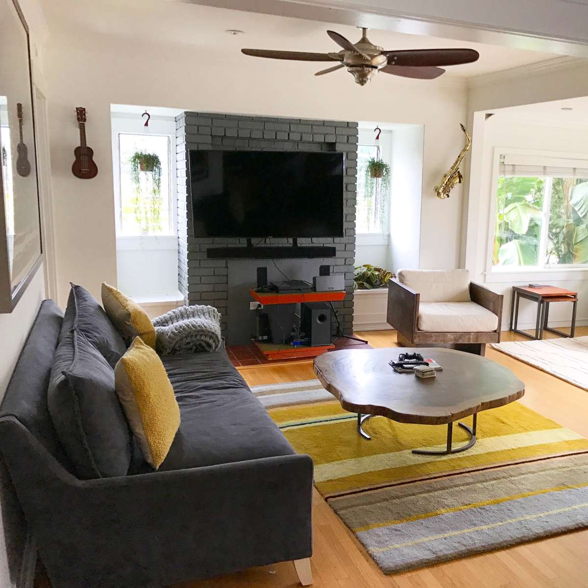 A modern living room featuring a gray sofa, two armchairs, a coffee table, a television on a black-brick wall, and windows with plants.