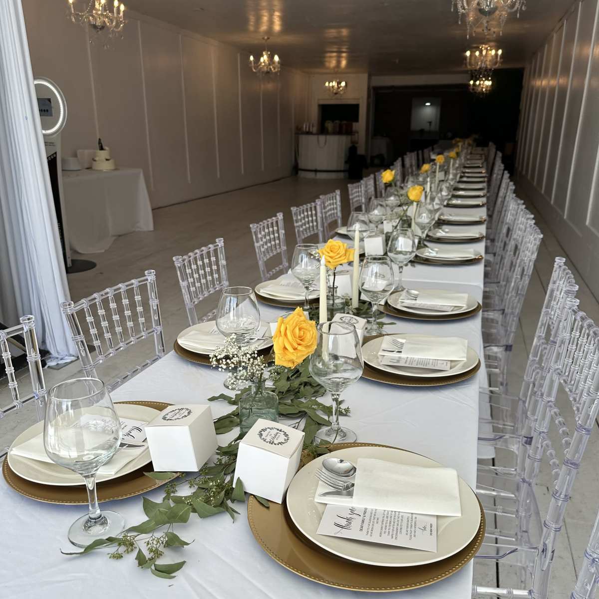 A long banquet table is elegantly set with yellow roses, glassware, and decorative items, arranged under chandeliers in a spacious room.