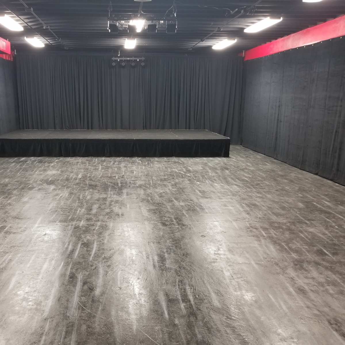 A dark, empty stage area with black curtains, a raised platform in the center, and a polished concrete floor.