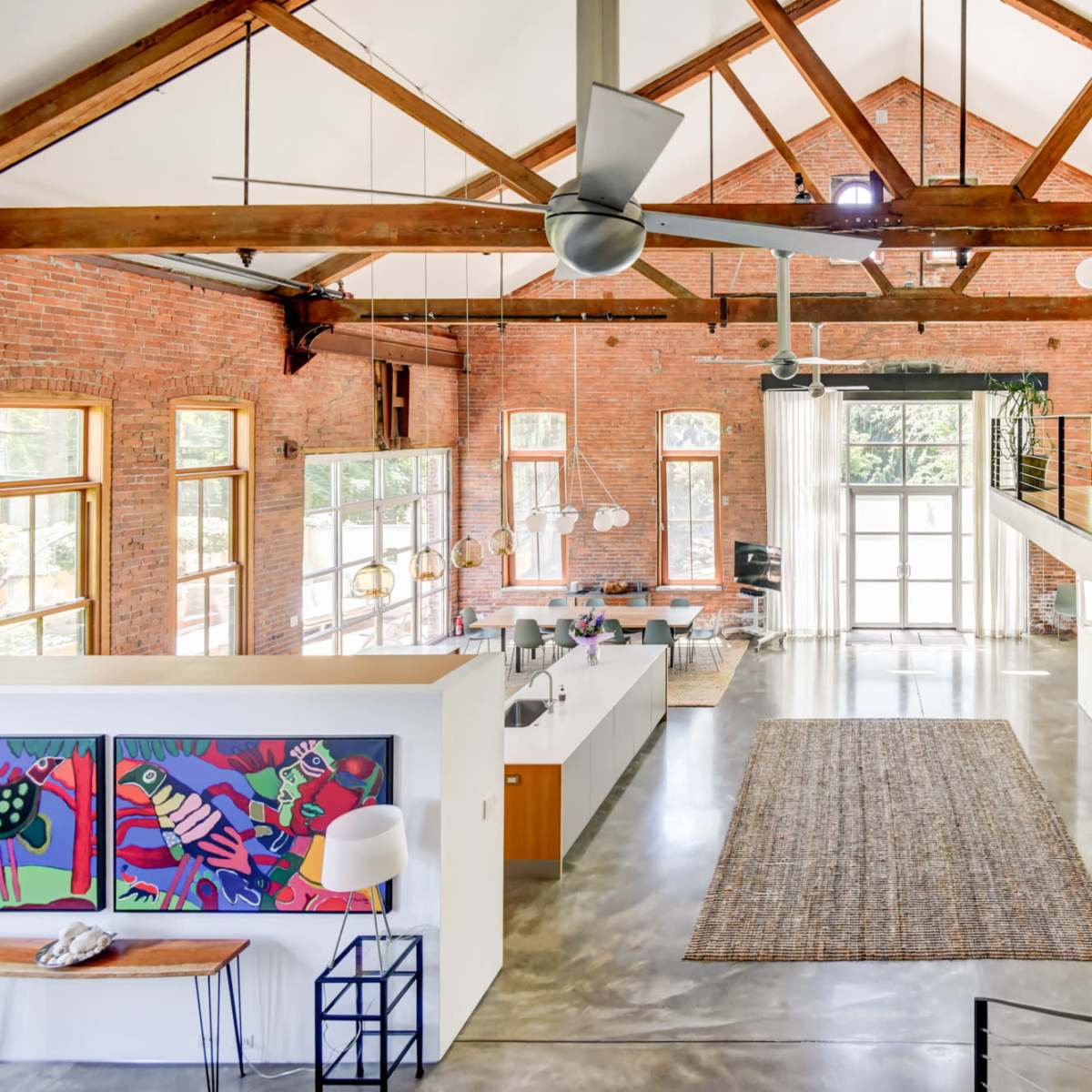 A spacious, modern interior featuring high ceilings with wooden beams, large windows, and a mix of brick and polished concrete surfaces.
