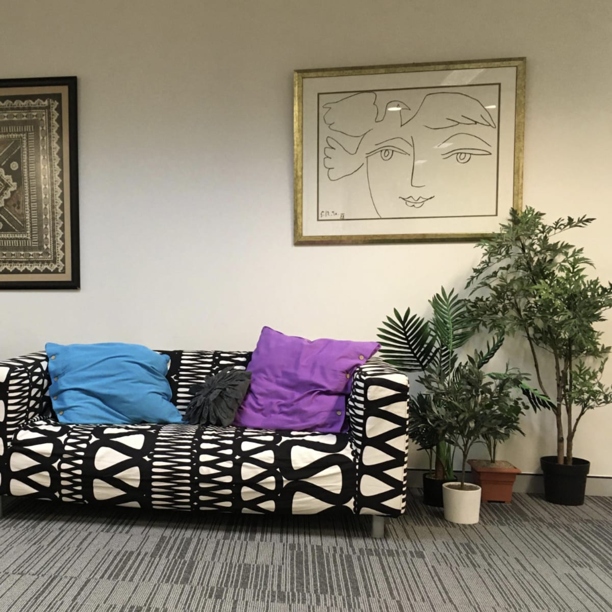 A living room corner with a patterned black and white couch adorned with blue and purple cushions, a black armchair, decorative wall art, and several potted plants.