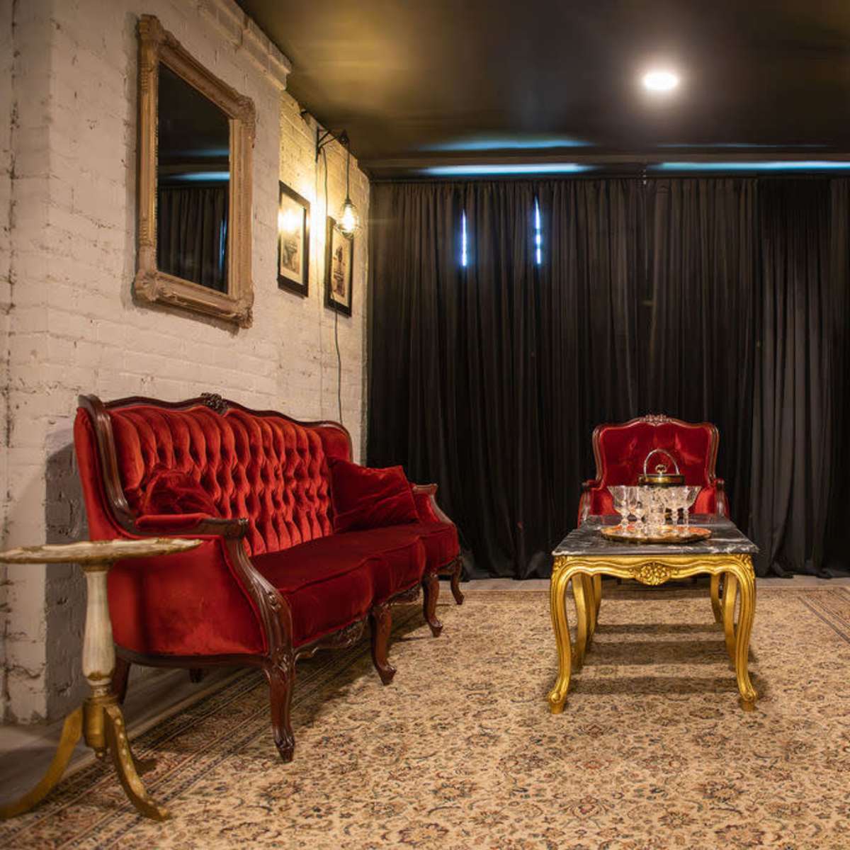 A sitting area with two red velvet sofas and a gold-accented coffee table on an ornate rug, surrounded by black curtains and framed mirrors on the walls.