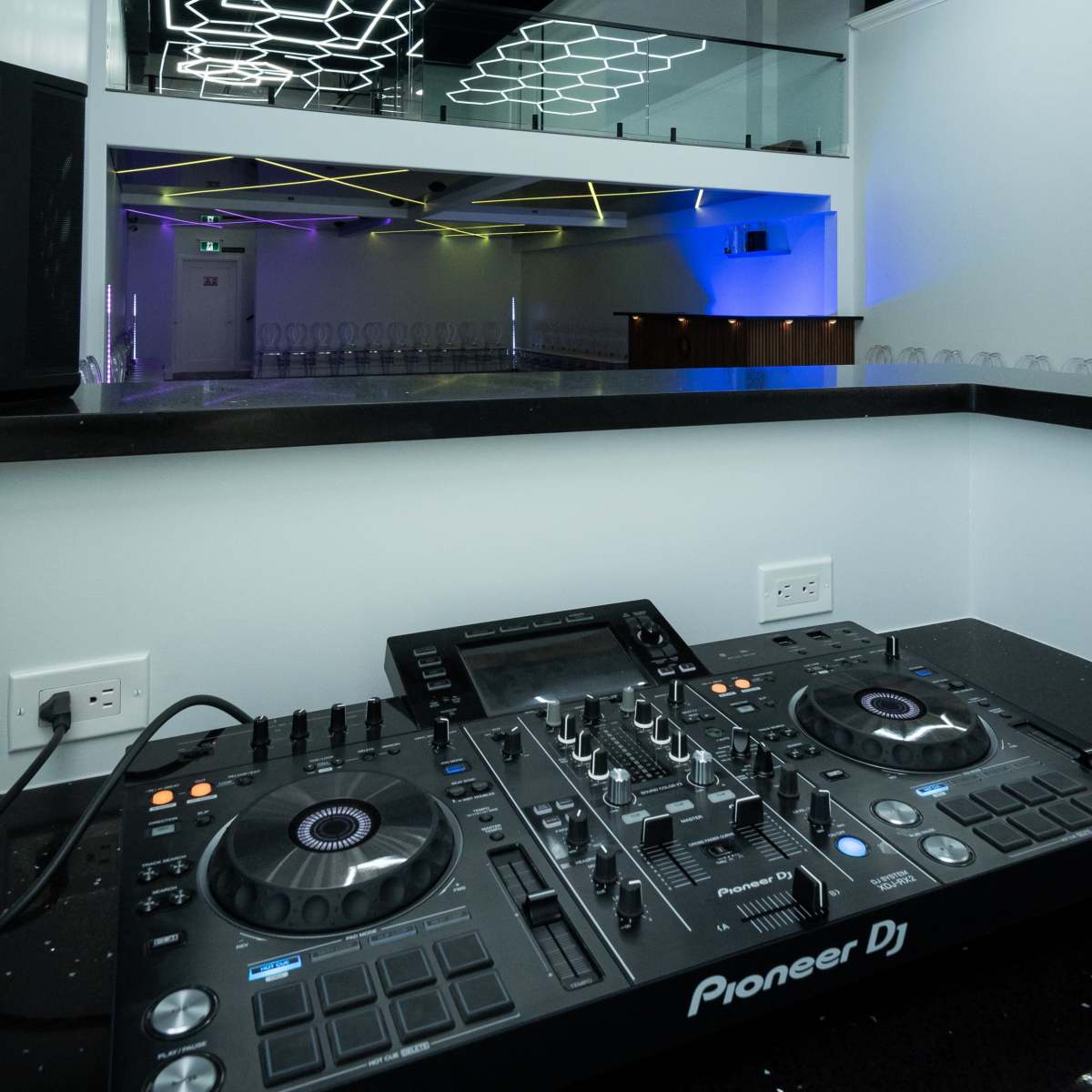 A modern DJ setup with a Pioneer DJ console in the foreground and a brightly lit, hexagon-patterned ceiling in an empty venue.