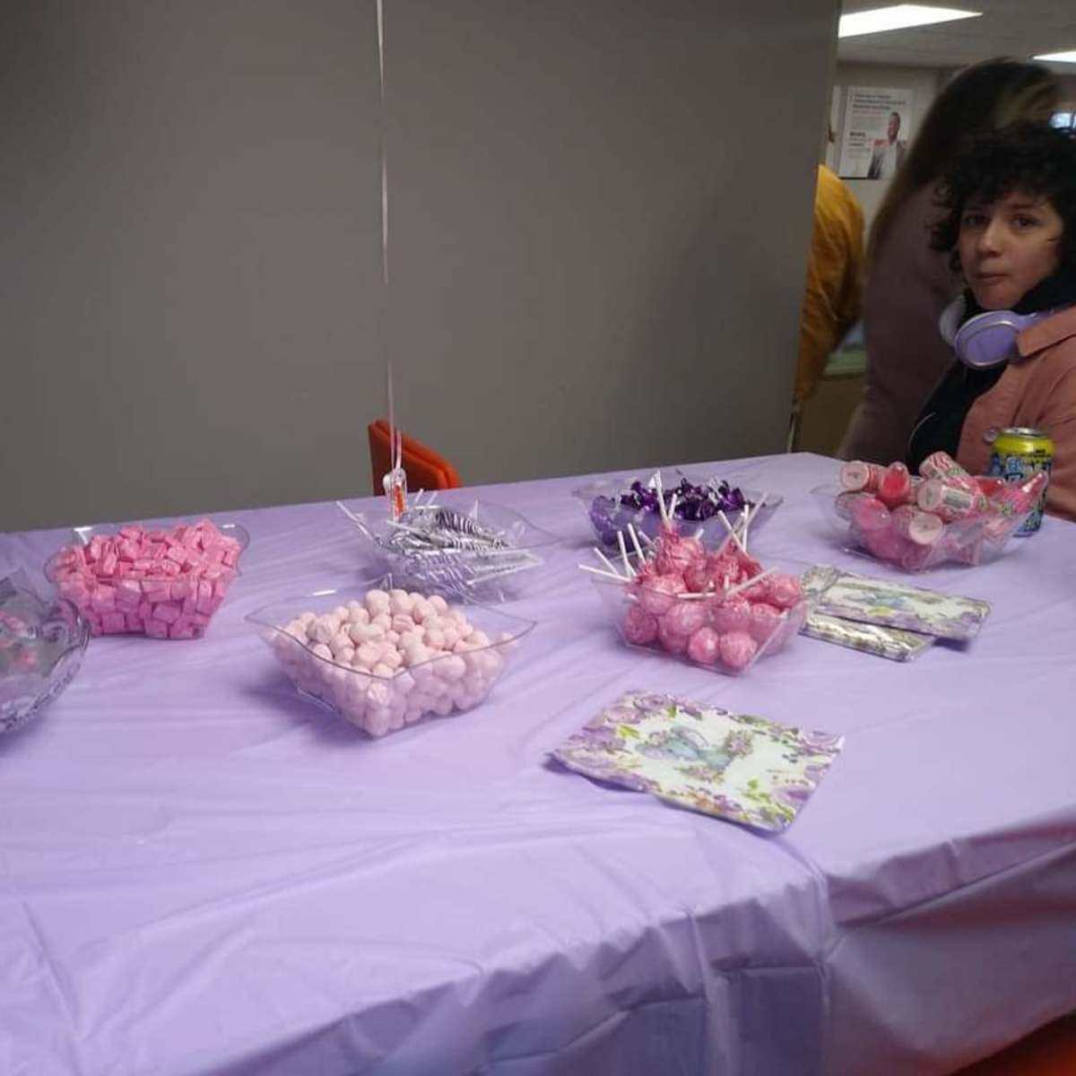A table covered with a purple tablecloth displays various candy dishes, including pink and purple sweets, beside a person sitting in a chair.