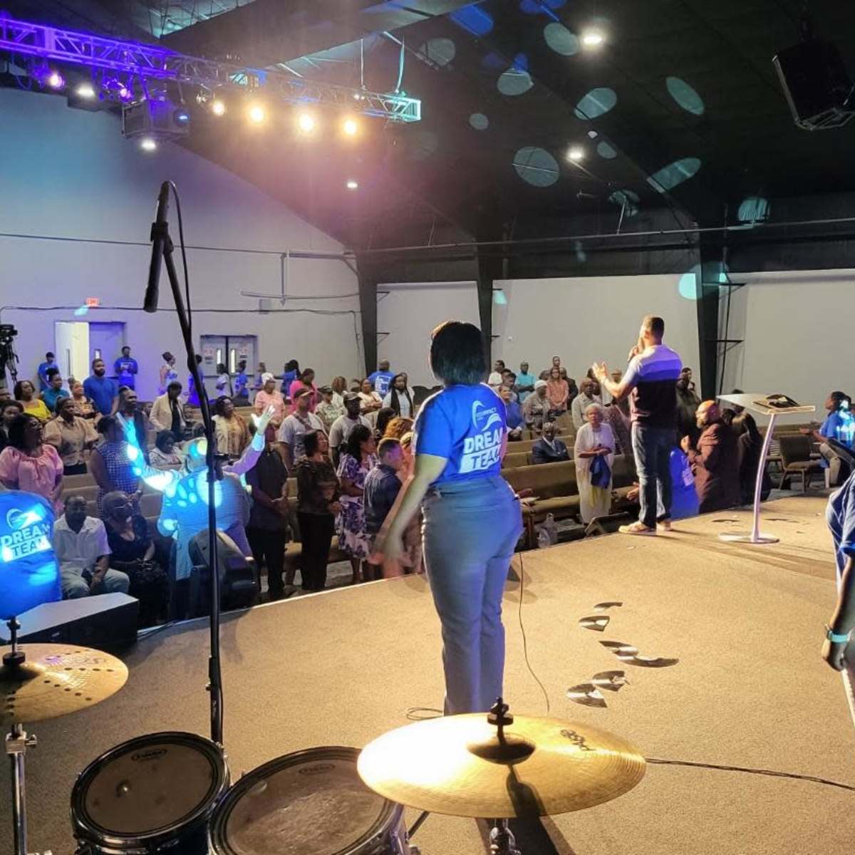 A group of people wearing matching blue shirts stands on a stage in a church, while an audience fills the pews in front of them.