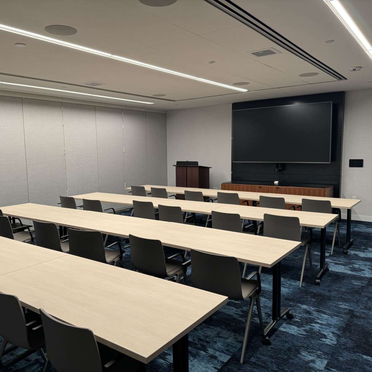 A modern conference room with several rows of tables and chairs arranged for a meeting, featuring a large screen at the front.
