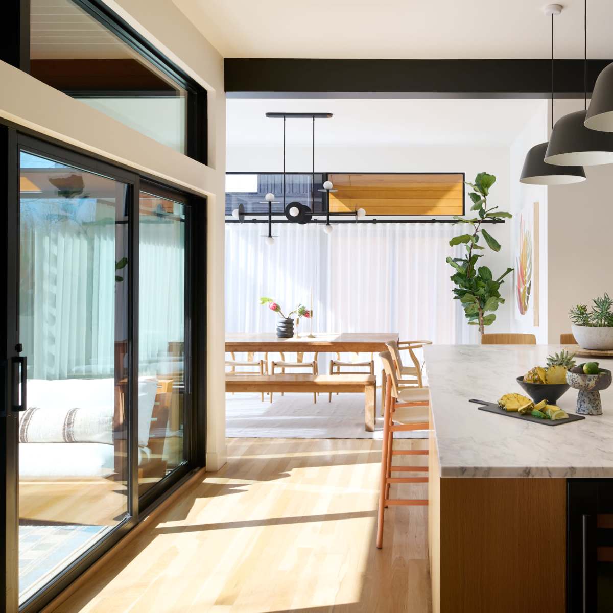 A modern kitchen open to a dining area, with a marble island, wooden dining table, and large windows allowing natural light.