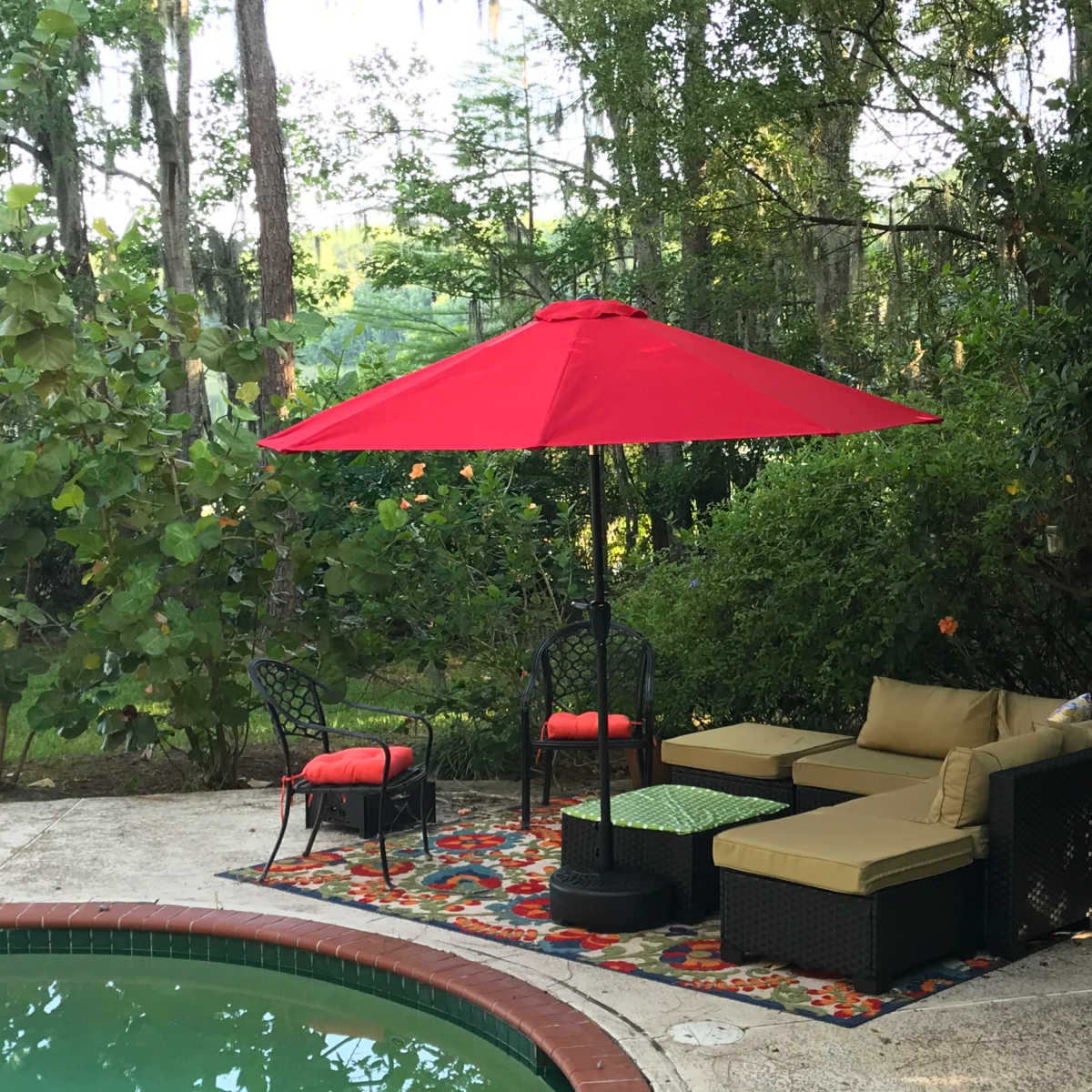 A red umbrella shades a seating area with a sofa and chairs beside a pool surrounded by greenery.