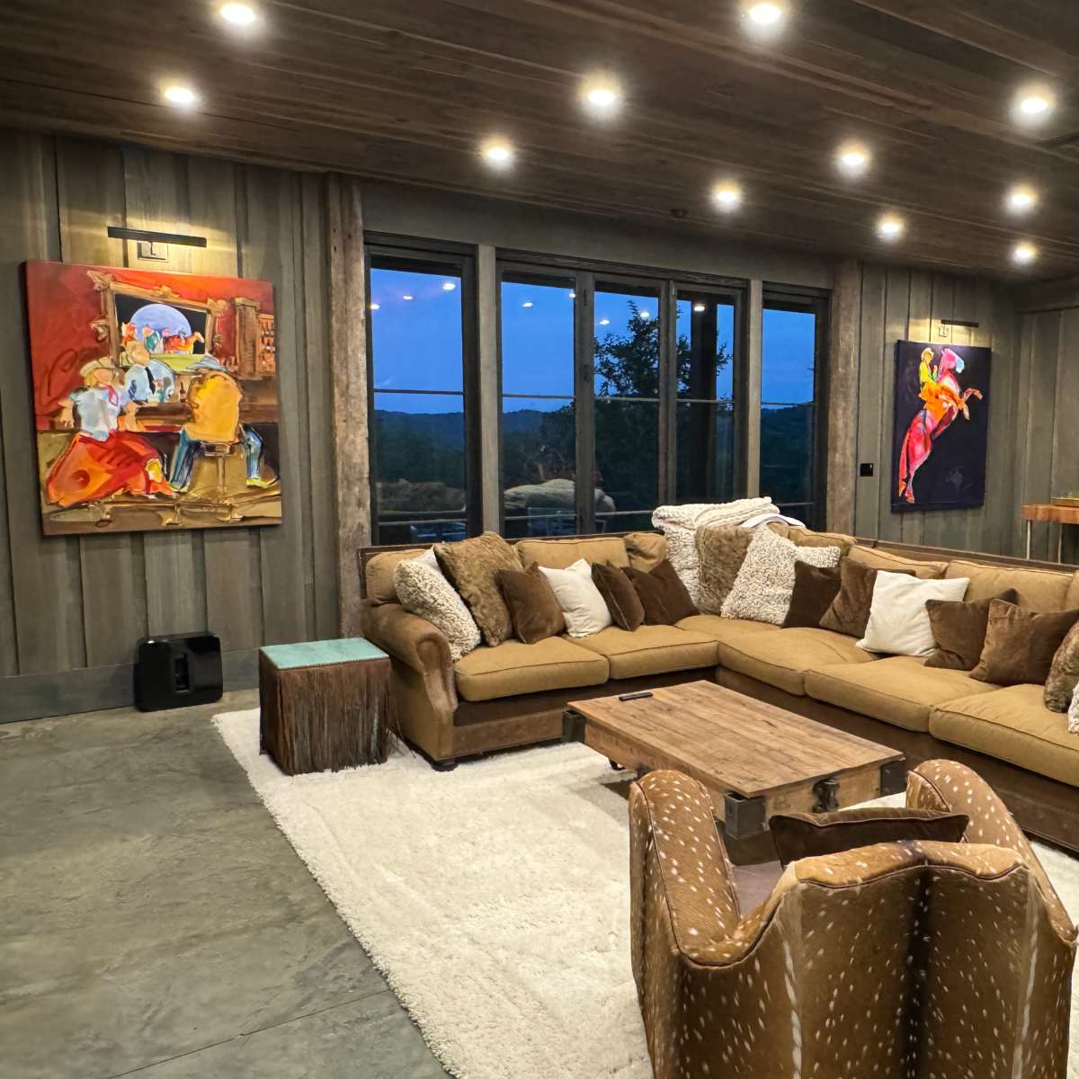 A spacious living room features a large sectional sofa with decorative pillows, a wooden coffee table, and two framed paintings on the walls, illuminated by recessed lighting.
