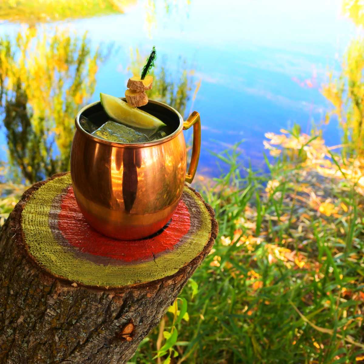 A copper mug filled with a drink and garnished with a lime slice sits on a painted log beside a body of water surrounded by greenery.