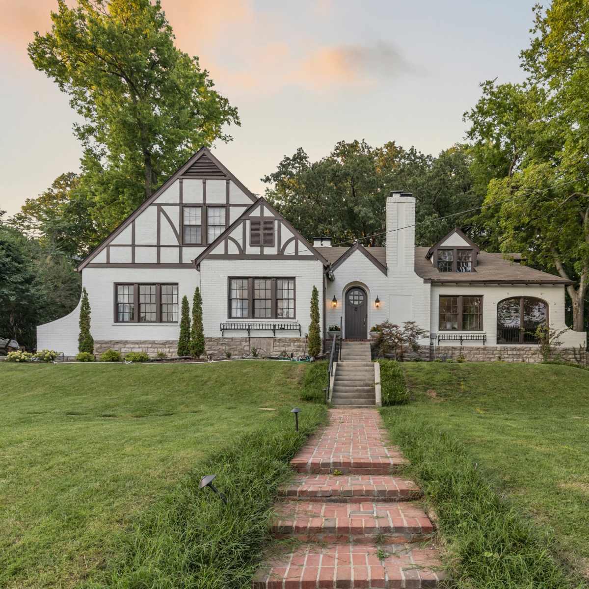 A two-story, Tudor-style house with a brick pathway leads to its front door, surrounded by green lawns and tall trees.