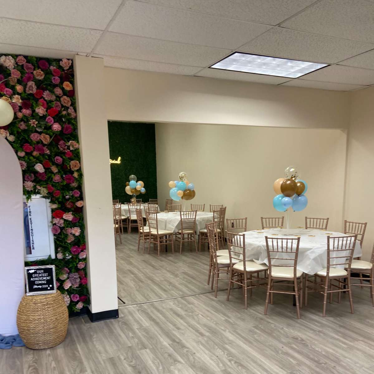 A decorated event space with wooden tables and chairs, a floral wall, and a mirrored area featuring balloons in blue and gold colors.