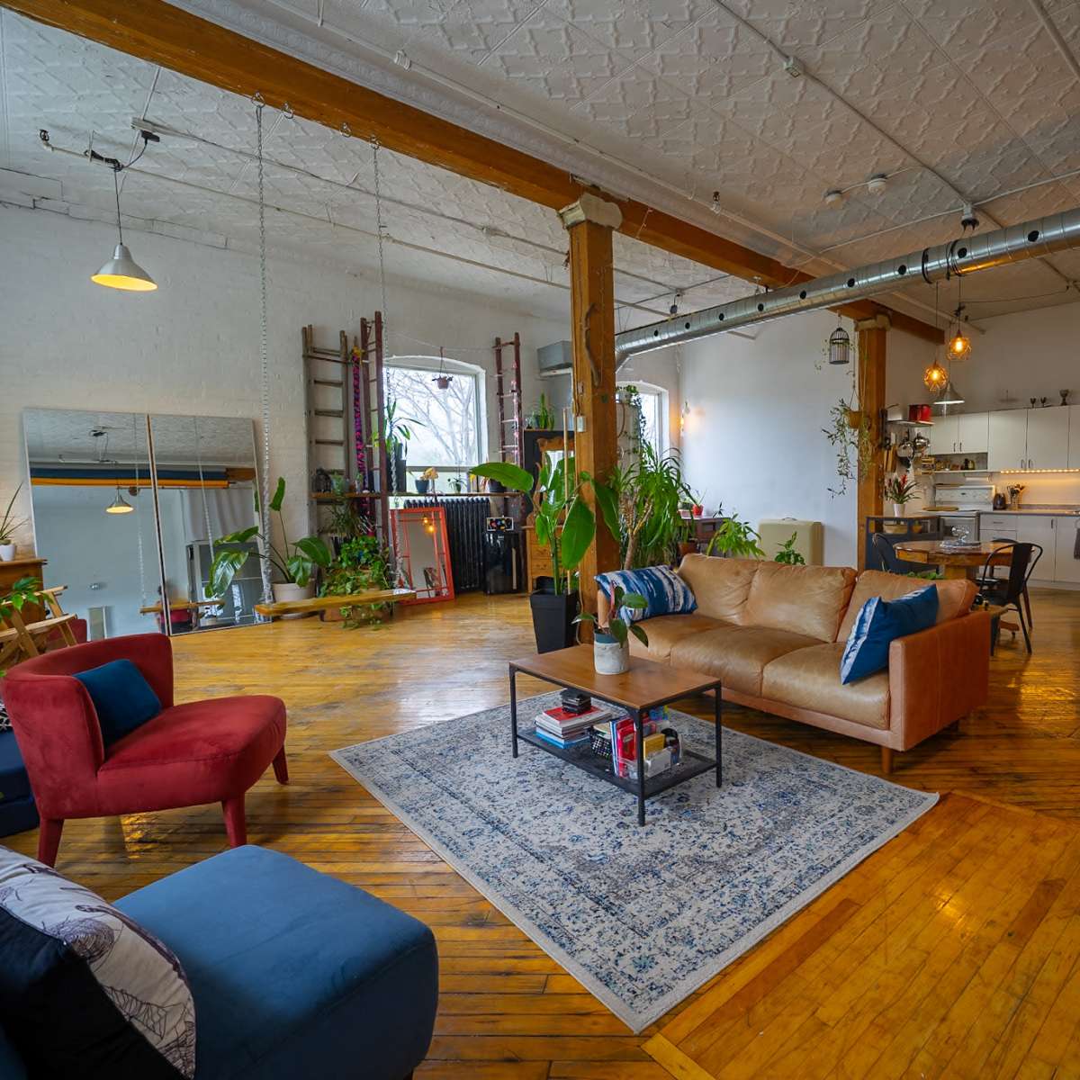 A spacious loft-style apartment with wooden floors, tall ceilings, and a mix of seating arrangements, including a brown couch and a red chair, surrounded by plants and decorative elements.