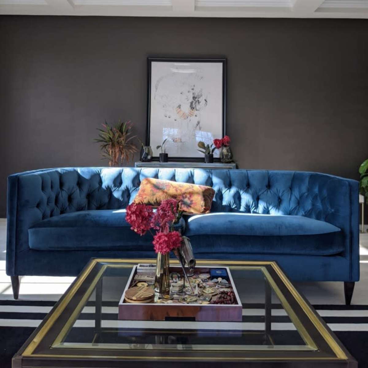 A blue velvet sofa is positioned against a dark wall, with a framed artwork above it and a glass coffee table adorned with decorative items and flowers in a vase.