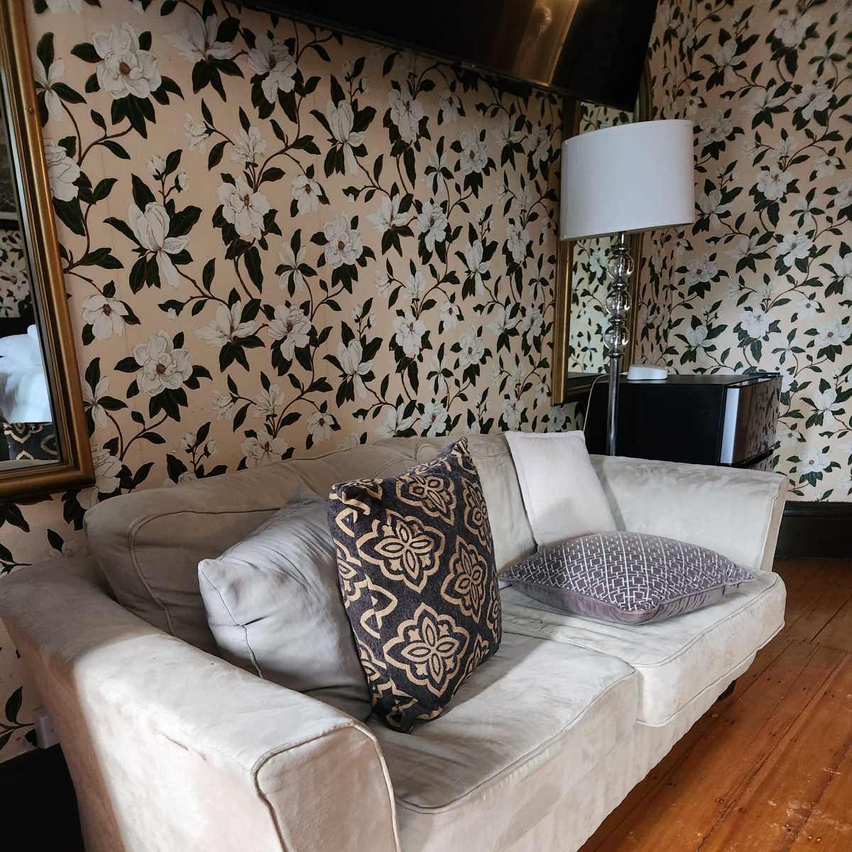 A light-colored sofa with decorative pillows in a room featuring floral wallpaper and a floor lamp.