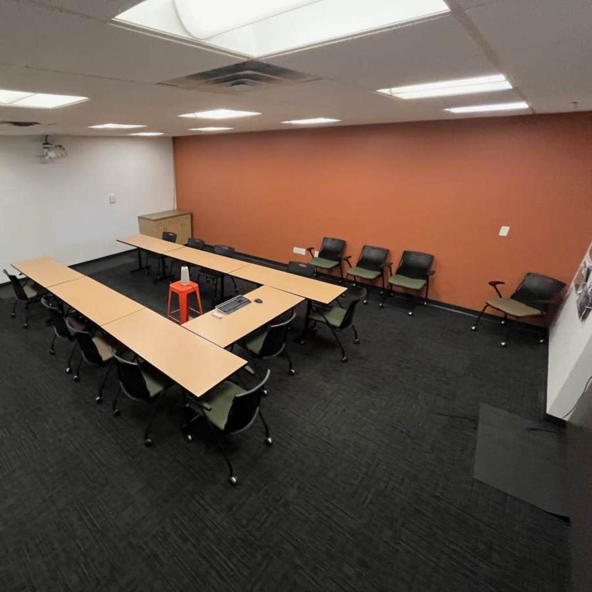 A large conference room features a rectangular table surrounded by black chairs, with a bright orange stool in the center and an orange accent wall.