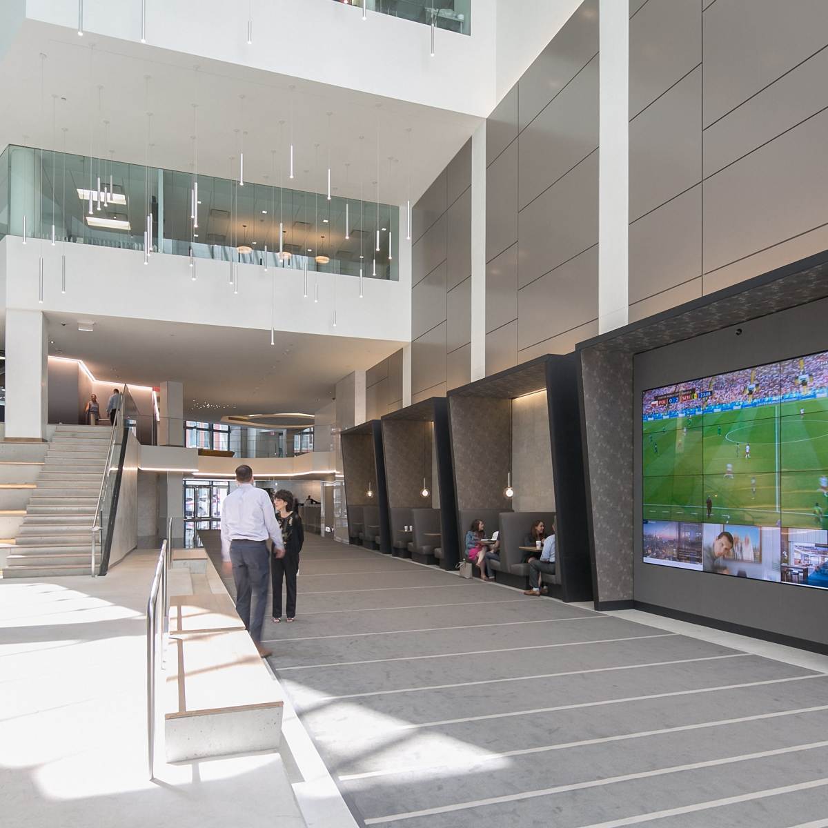 A modern, open atrium with a staircase leading to an upper level, featuring large screens displaying a sports event along one wall.