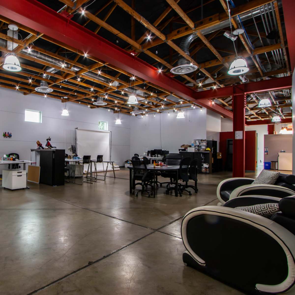 A modern office space with open areas, multiple workstations, and colorful seating arrangements under a well-lit ceiling with exposed beams.