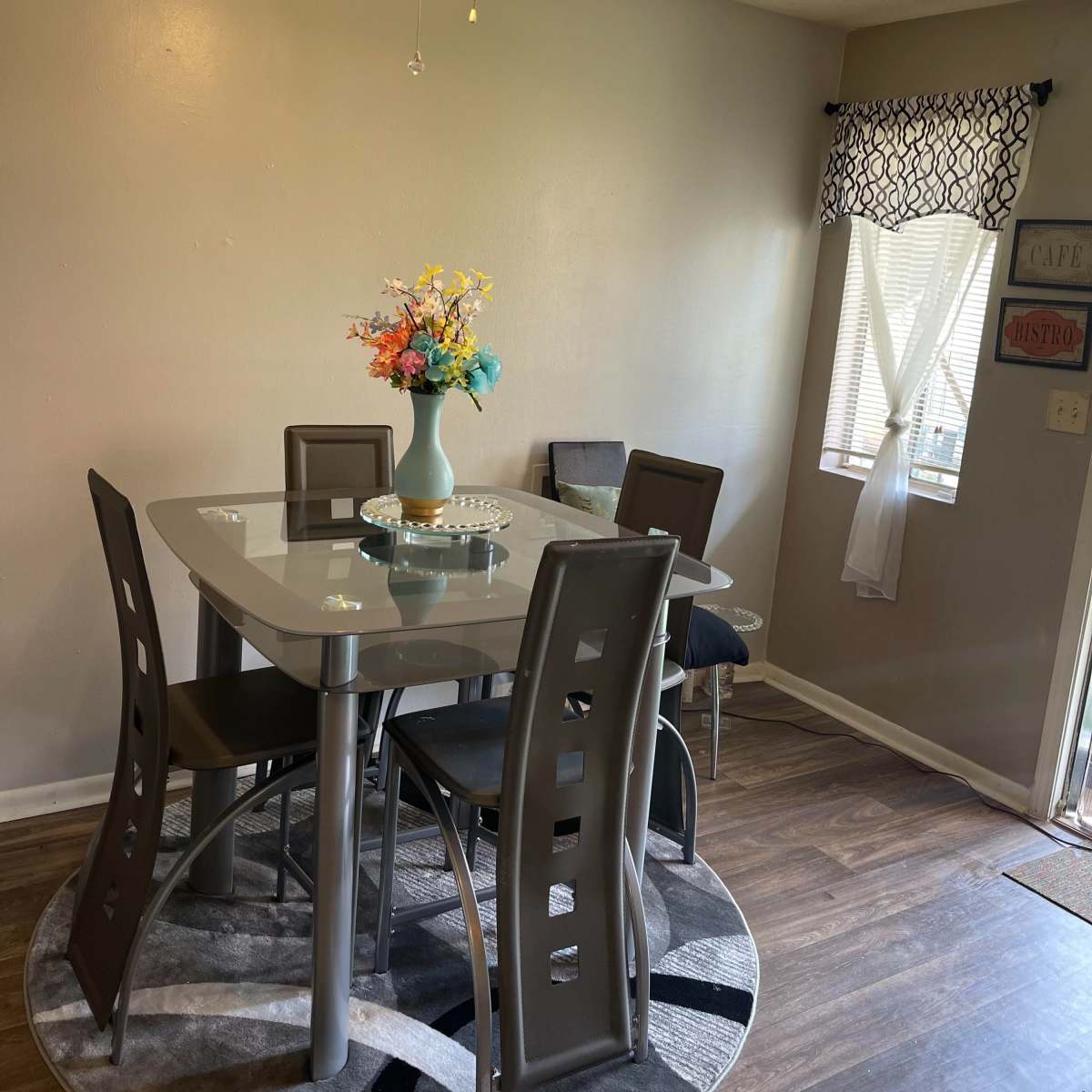 A modern dining area features a glass-top table surrounded by four chairs, a decorative vase with flowers, and a circular rug under the table.