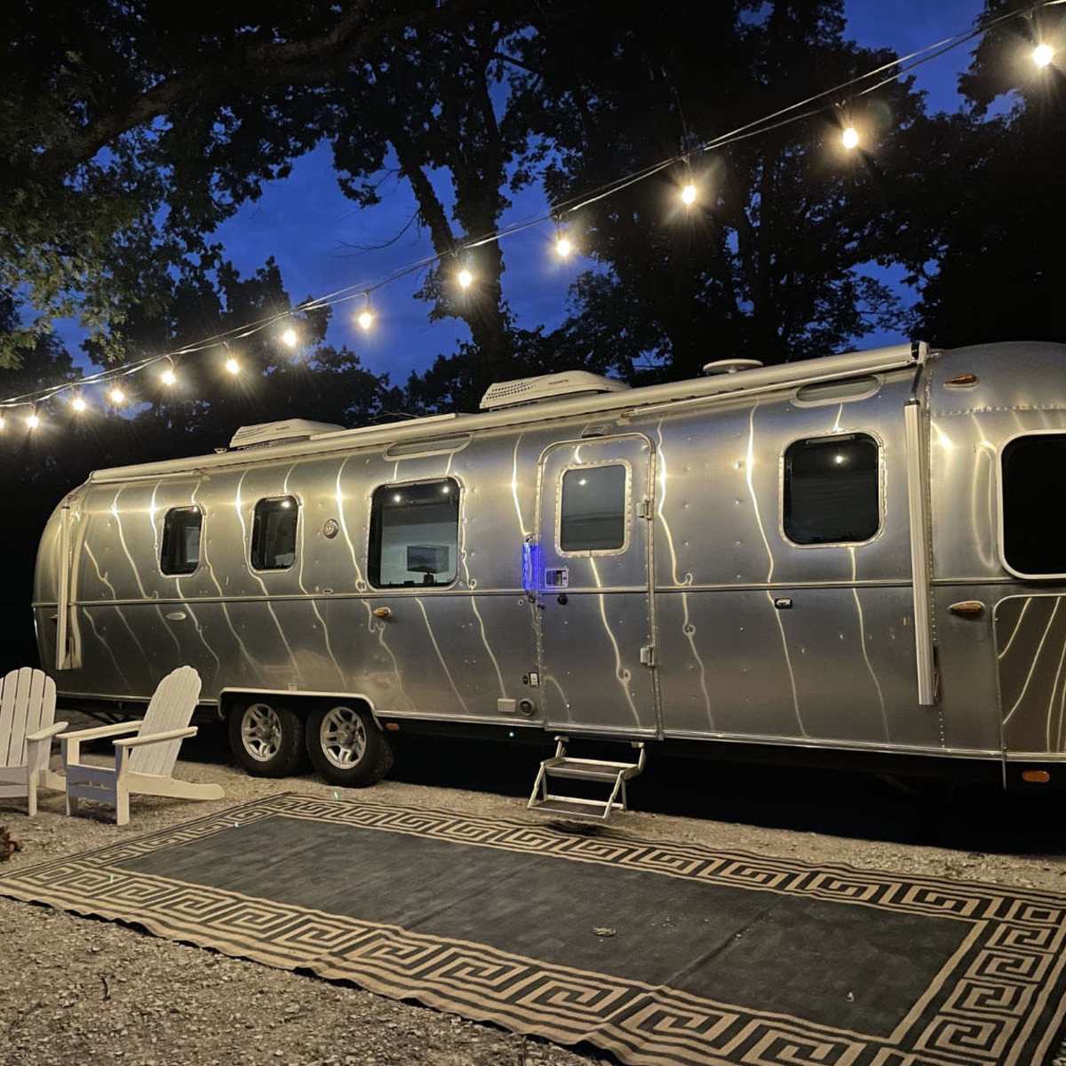 A polished silver Airstream trailer is parked under string lights next to a fire pit and two Adirondack chairs on a patterned rug.