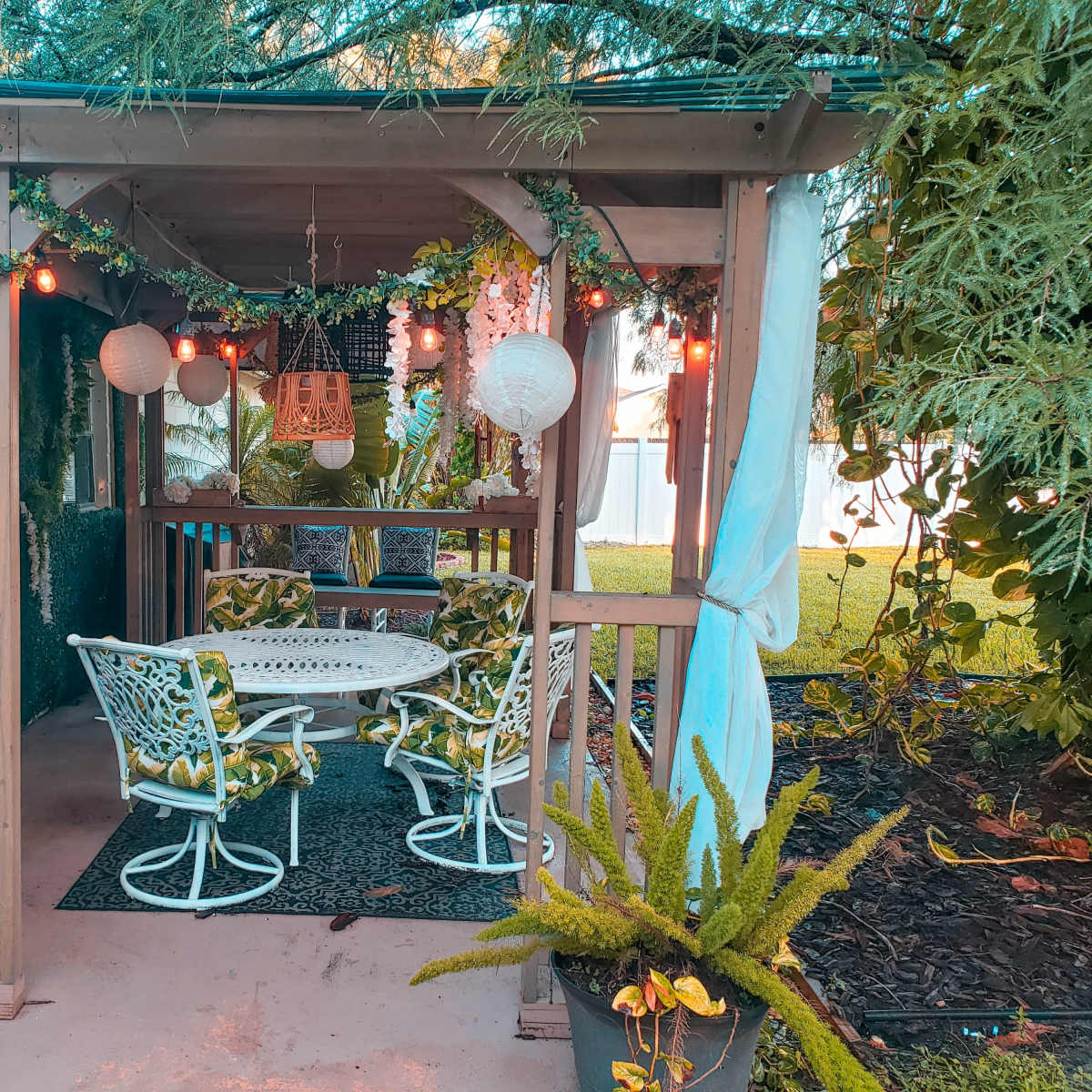 A cozy outdoor seating area, featuring a round table and four chairs surrounded by greenery and decorative lanterns.