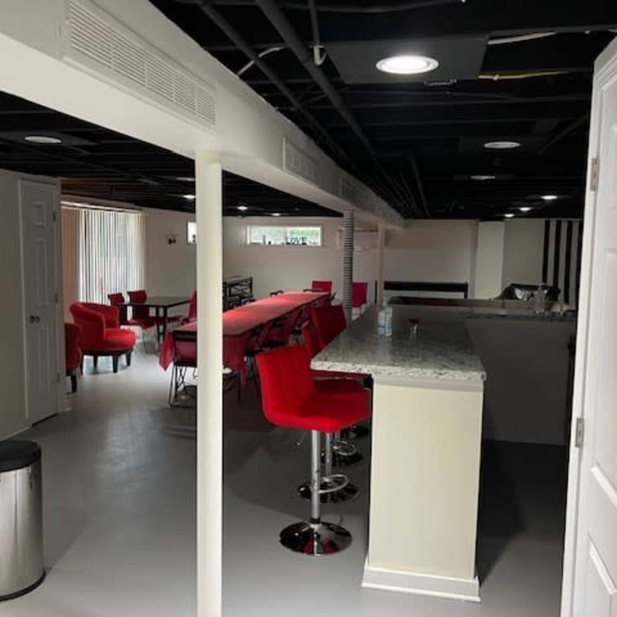 A spacious basement area with a bar counter, red stools, and a long dining table surrounded by red chairs.