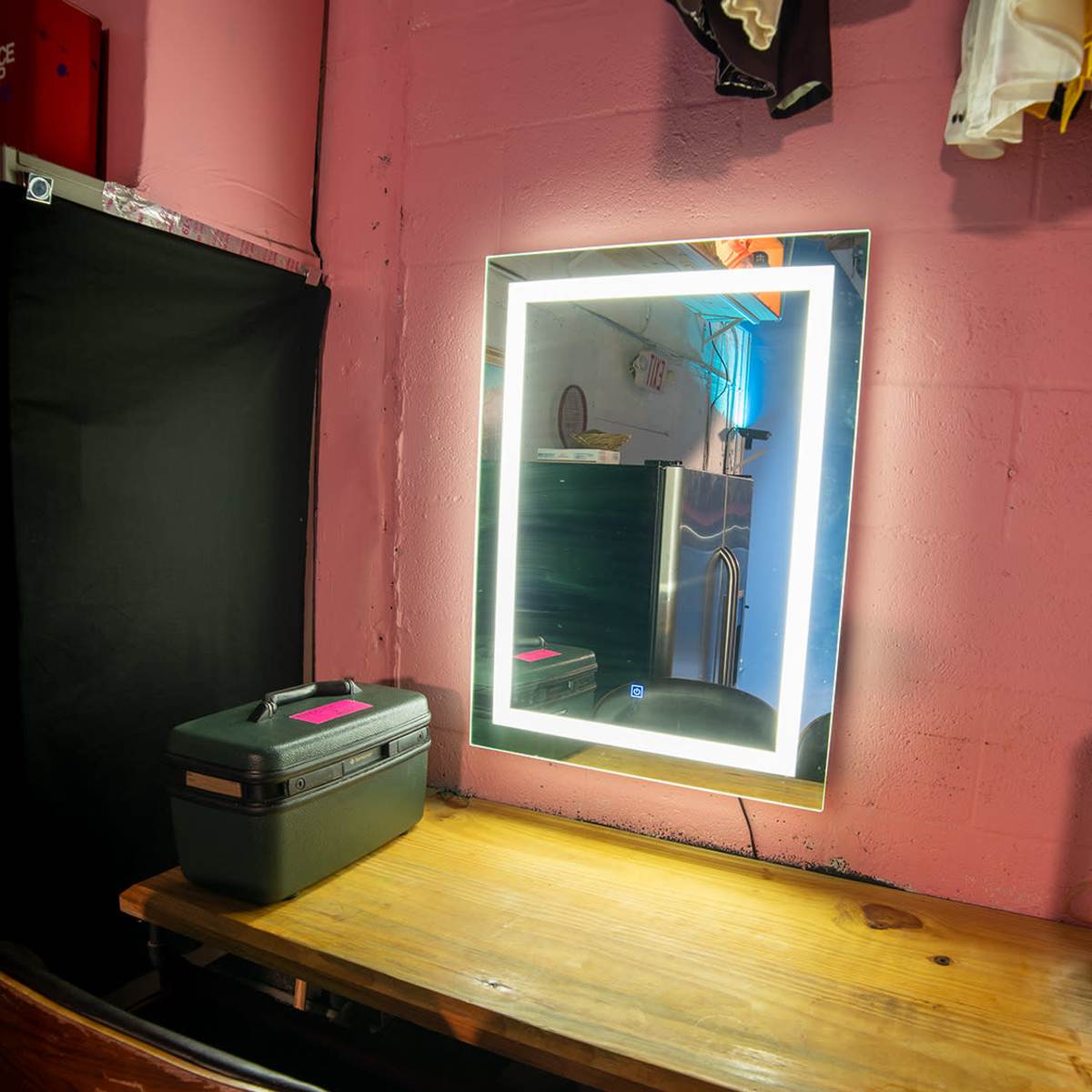 A wooden table sits against a pink wall, featuring a backlit mirror and a black storage box nearby.