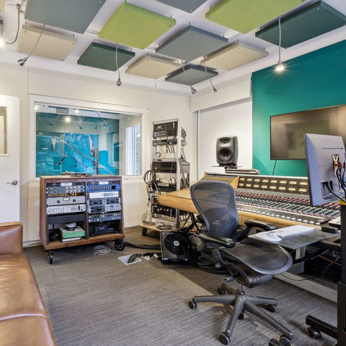 A recording studio with a mixing console, various audio equipment, a computer workstation, and a lounge area with a brown sofa.