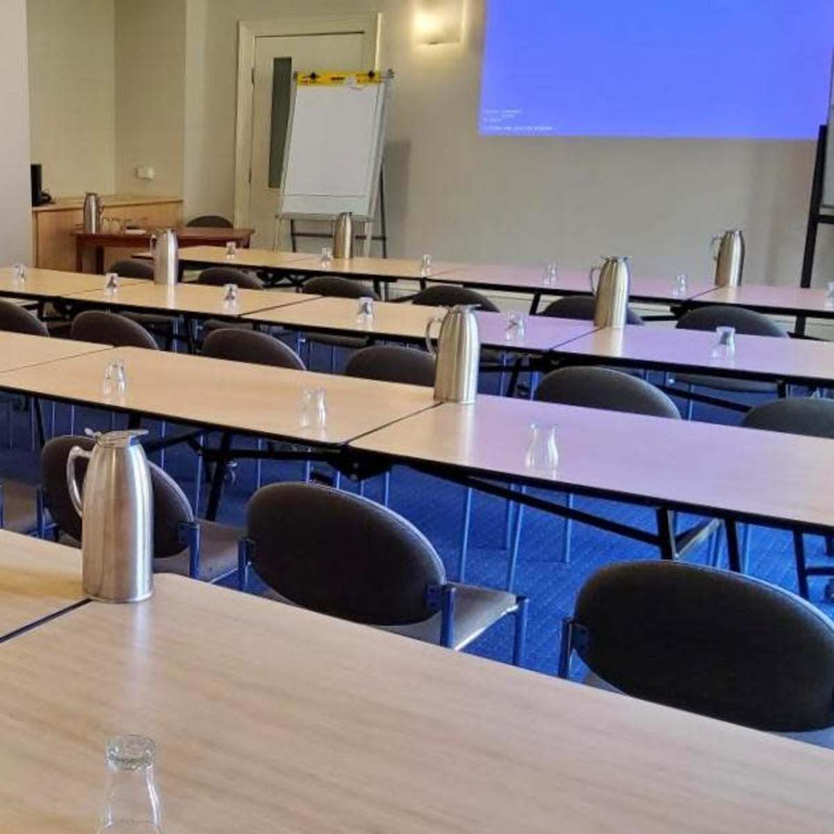 A conference room set up with multiple tables arranged in a U-shape, each equipped with water pitchers and small glasses.