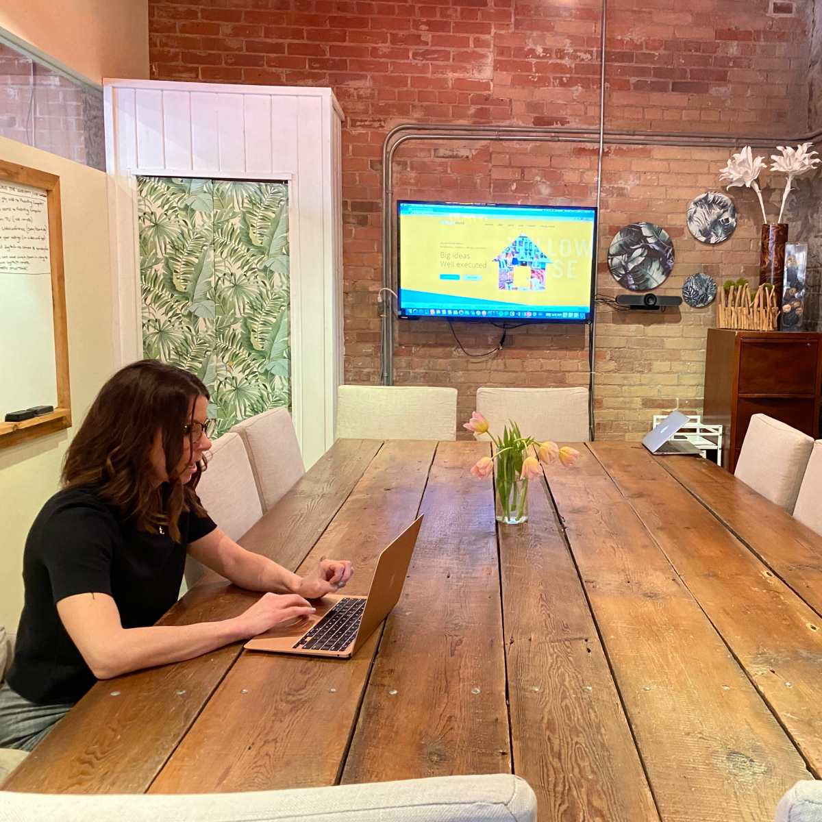 A person is working on a laptop at a long wooden table in a meeting room with a large screen displaying a colorful website in the background.