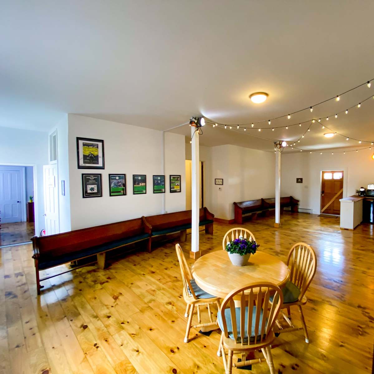 A spacious, well-lit interior featuring a dining area with a round table and chairs, wooden floors, and a kitchenette in the background.