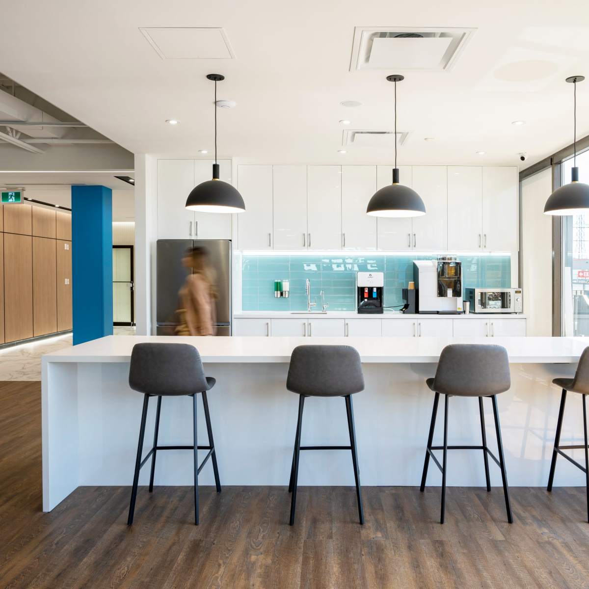 A modern kitchen features a long white countertop with four bar stools and sleek cabinetry against a blue backsplash.