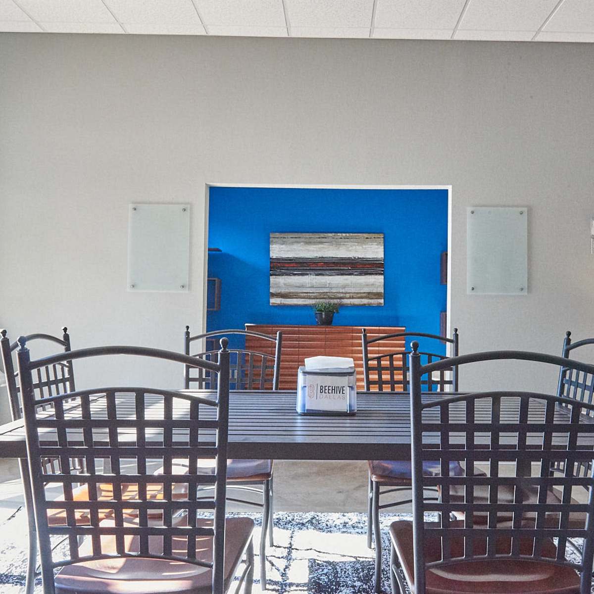 A spacious dining area features a metal table surrounded by wooden chairs, with a blue wall and an abstract painting in the background.