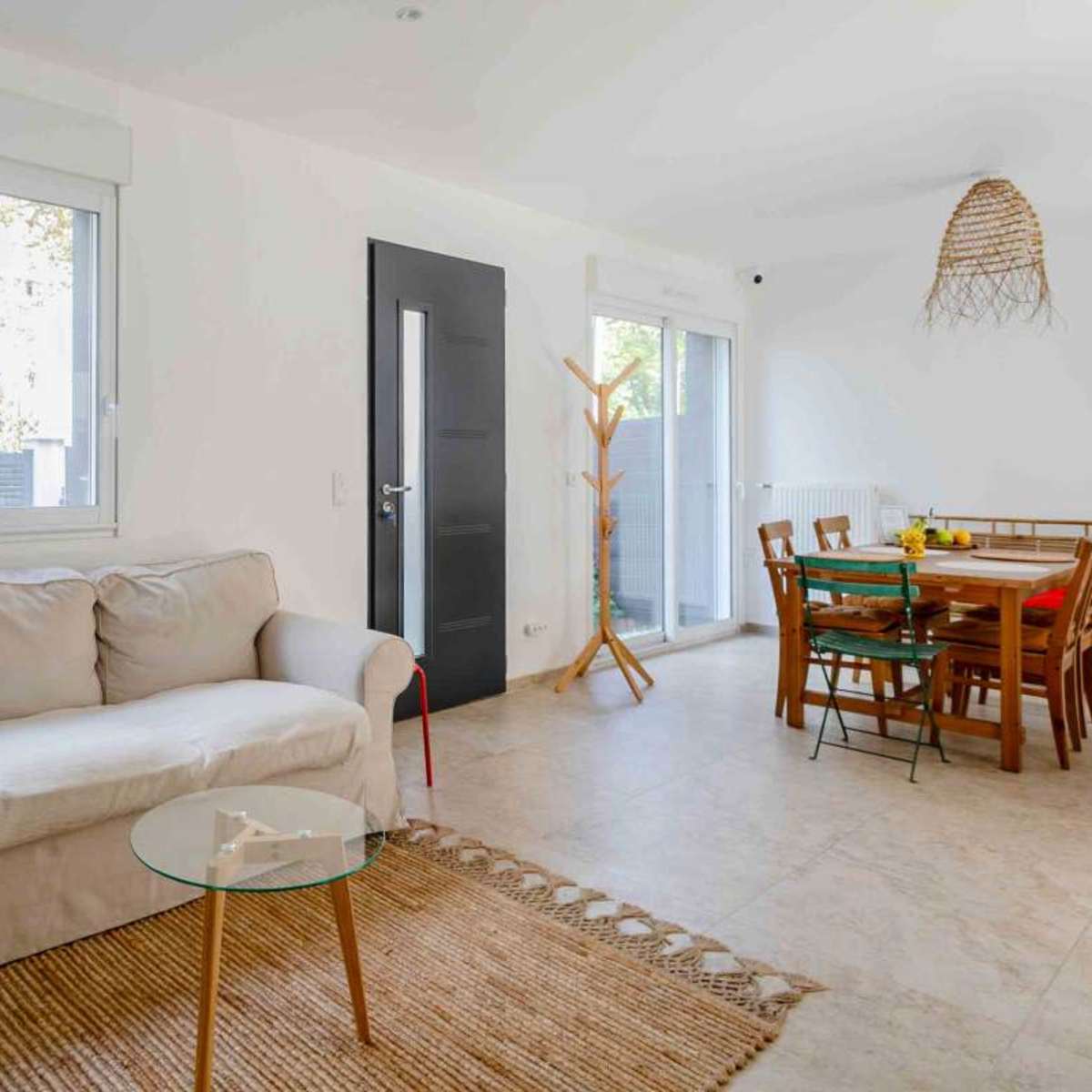 A light-filled living space features a beige couch, a glass coffee table, and a wooden dining table surrounded by chairs.