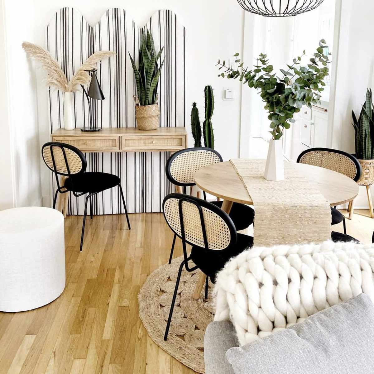 A dining area features a round table surrounded by black and rattan chairs, with a light fixture overhead and various indoor plants in decorative pots.