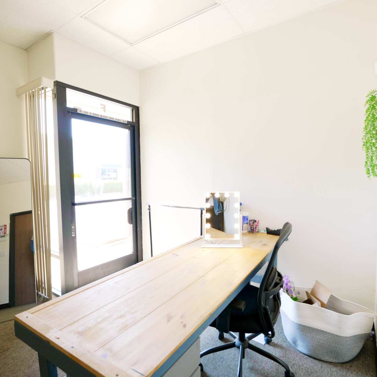 A desk sits in a bright, minimalist office space with large windows, and a potted plant is visible on the wall.