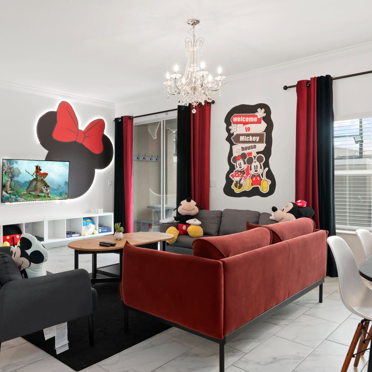 The living room features a mix of red and gray furniture, a wall decorated with Mickey Mouse art, and a chandelier above a seating area.