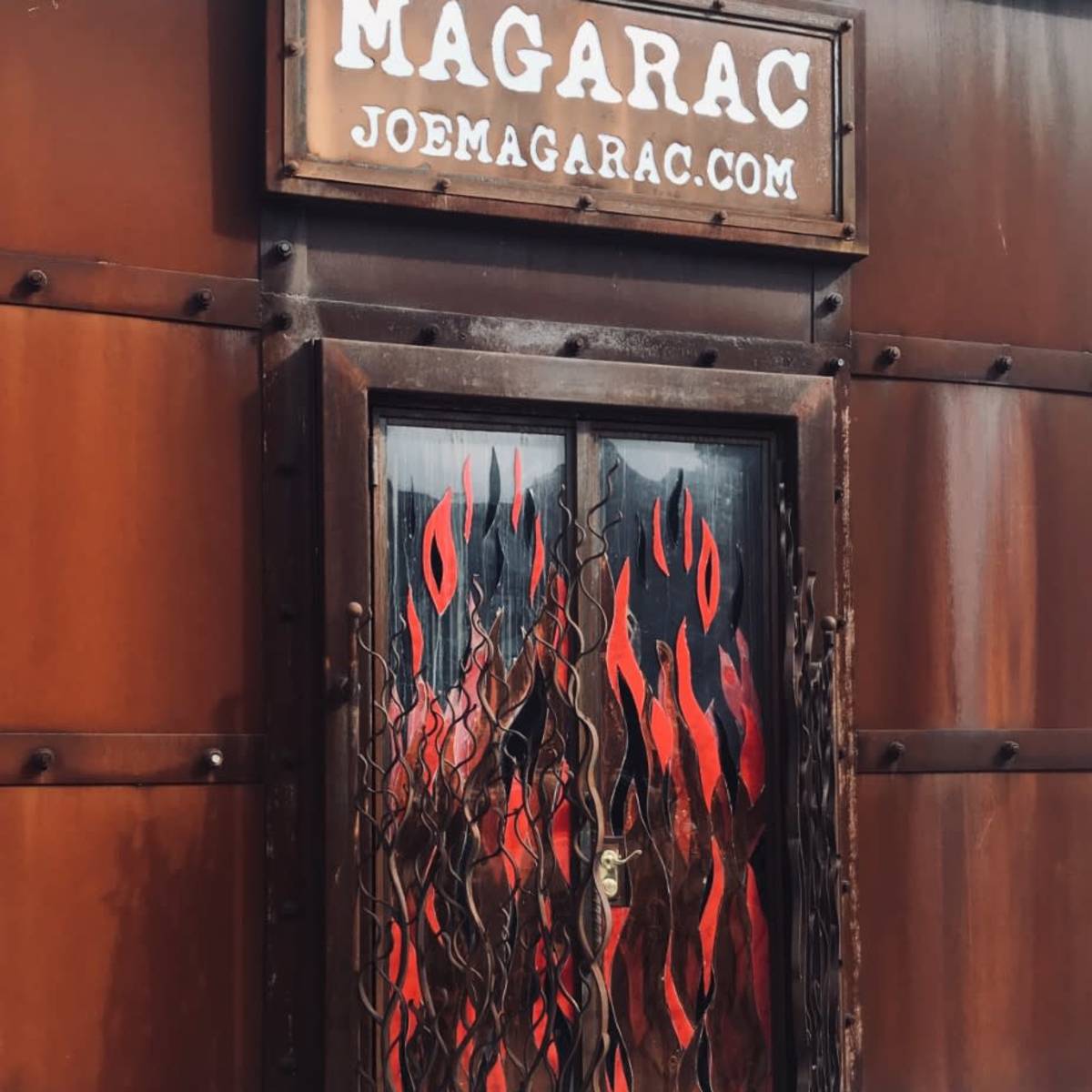A metal door with a flame-themed design behind a rusted metal exterior and a sign overhead that reads "MAGARAC" along with the website "JOEMAGARAC.COM."