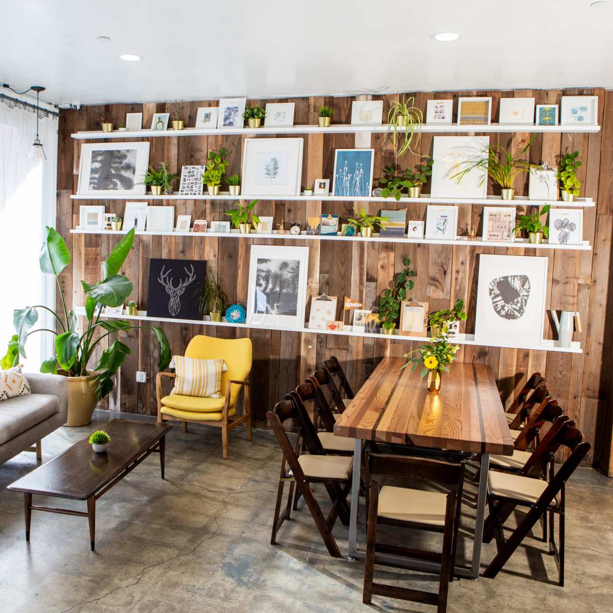 A cozy interior space featuring a wooden wall with shelves displaying framed art and plants, alongside a gray couch, a yellow chair, and a dining table with chairs.