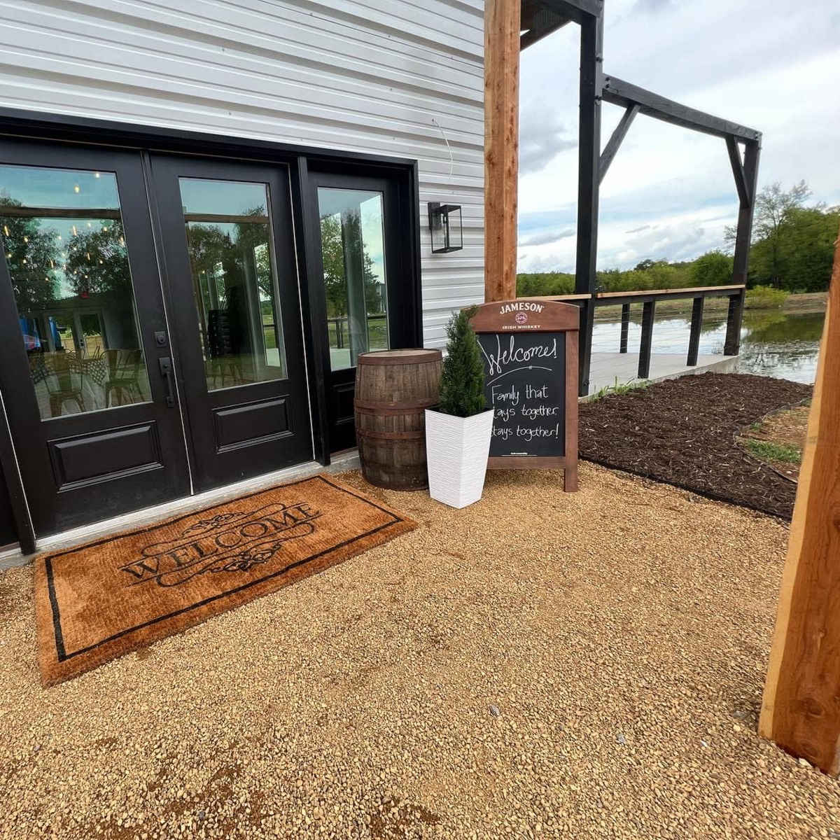 The entrance features a welcome mat in front of double black doors, flanked by two wooden barrels and a chalkboard sign beside a pond.