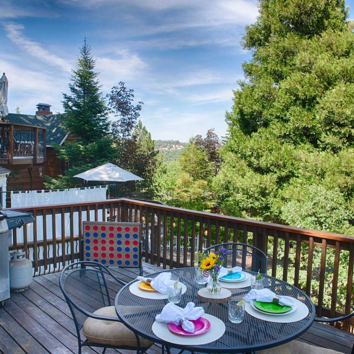A wooden deck with a table set for dining, featuring colorful plates and a flower centerpiece, alongside a barbecue grill and a view of trees in the background.