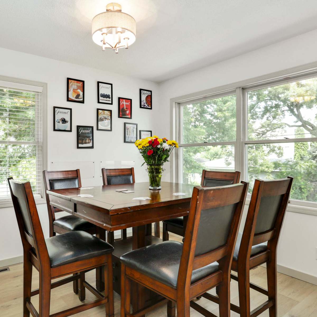 A dining area features a wooden table surrounded by eight chairs, with framed pictures on the walls and a bouquet of flowers on the table.