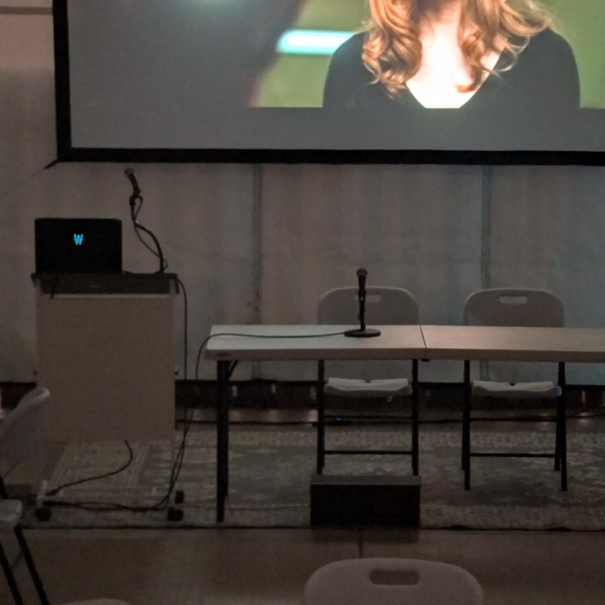 A microphone stands on a table in front of a projected screen displaying a person's face, with empty chairs arranged behind.