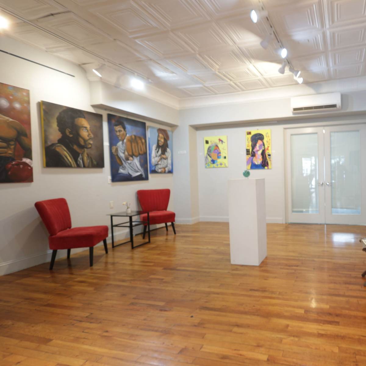 The gallery features an arrangement of paintings on the walls, with two seating areas and a central display pedestal in a bright, wooden-floored space.