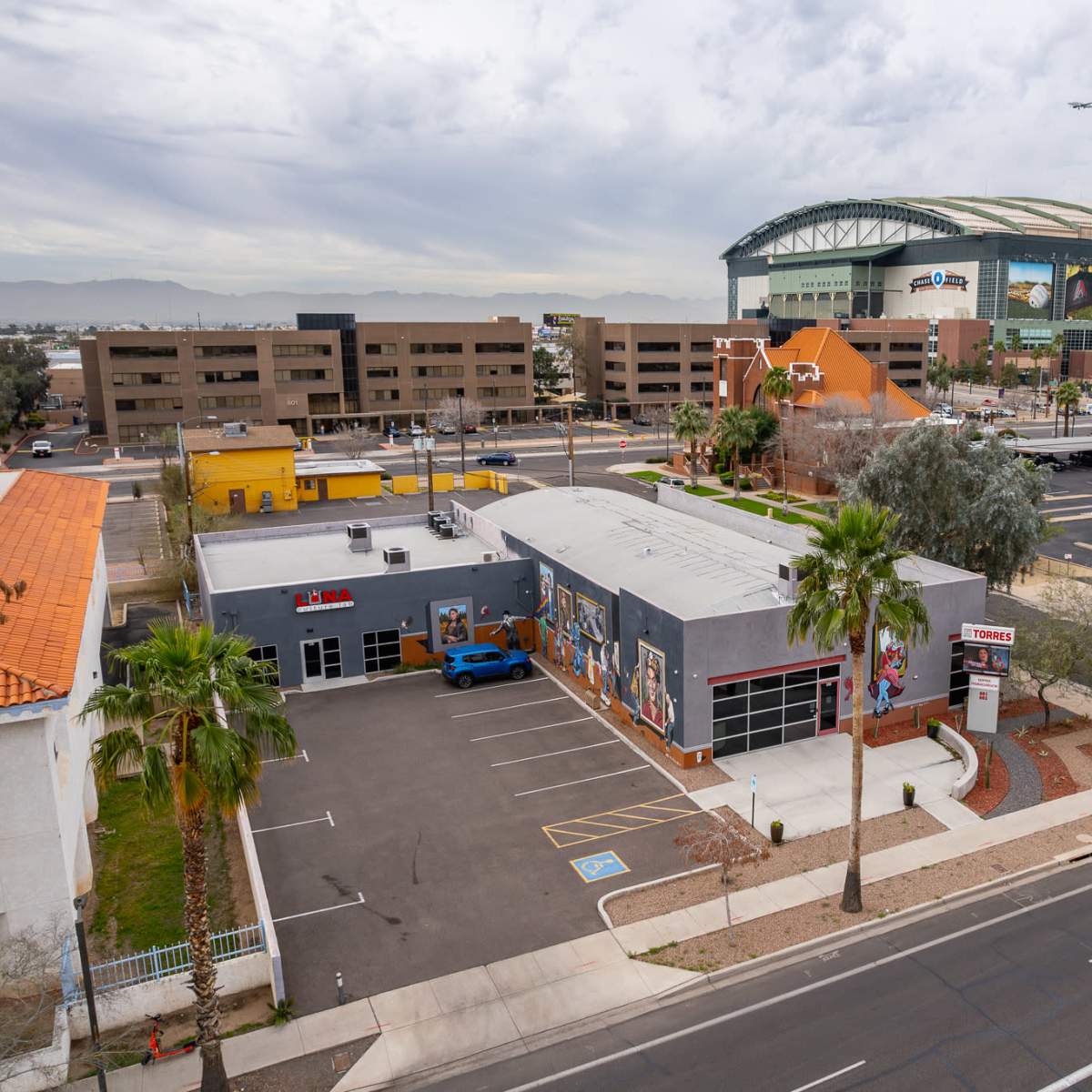 A view of a commercial area featuring a building with colorful murals, palm trees, and a large sports arena in the background.