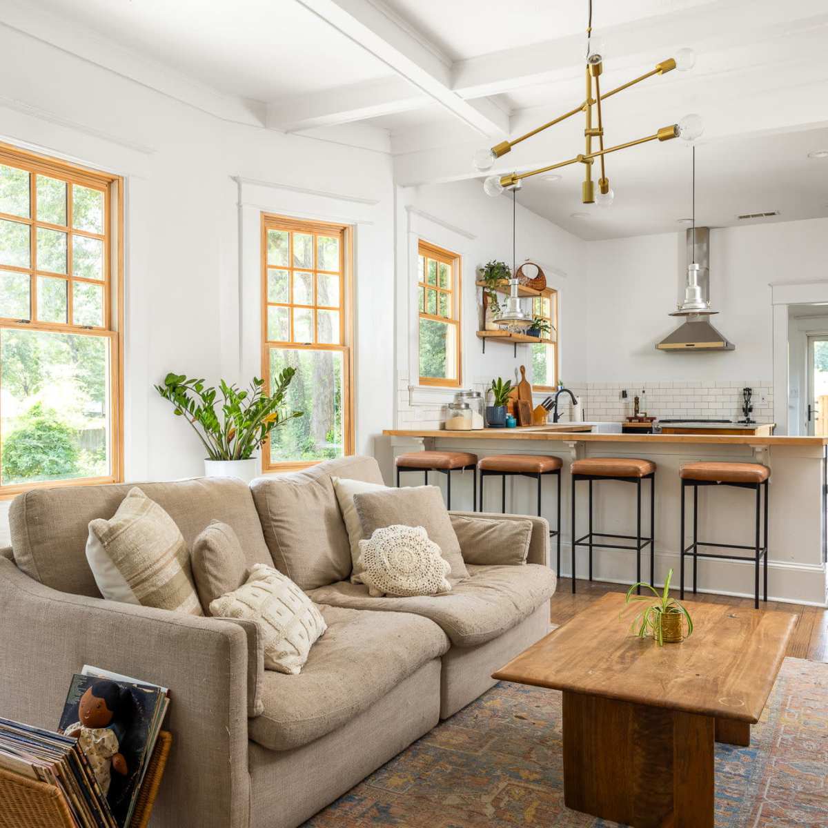 A cozy living room features a beige sofa and a wooden coffee table, with large windows allowing natural light to illuminate the space and a kitchen visible in the background.