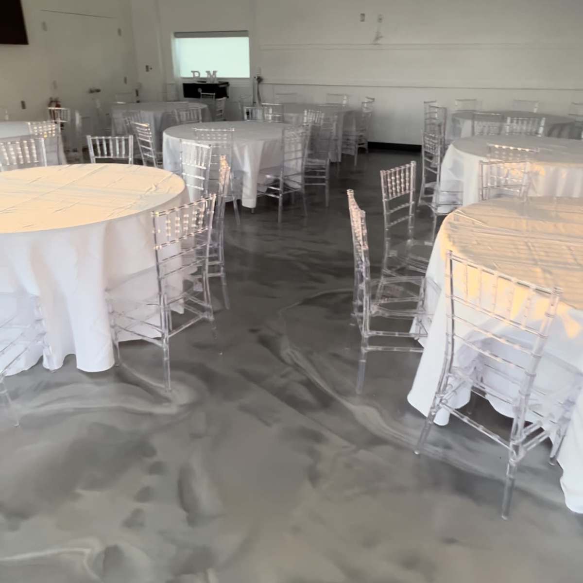 A spacious event area with several round tables covered in white tablecloths and clear acrylic chairs arranged on a marbled gray floor.