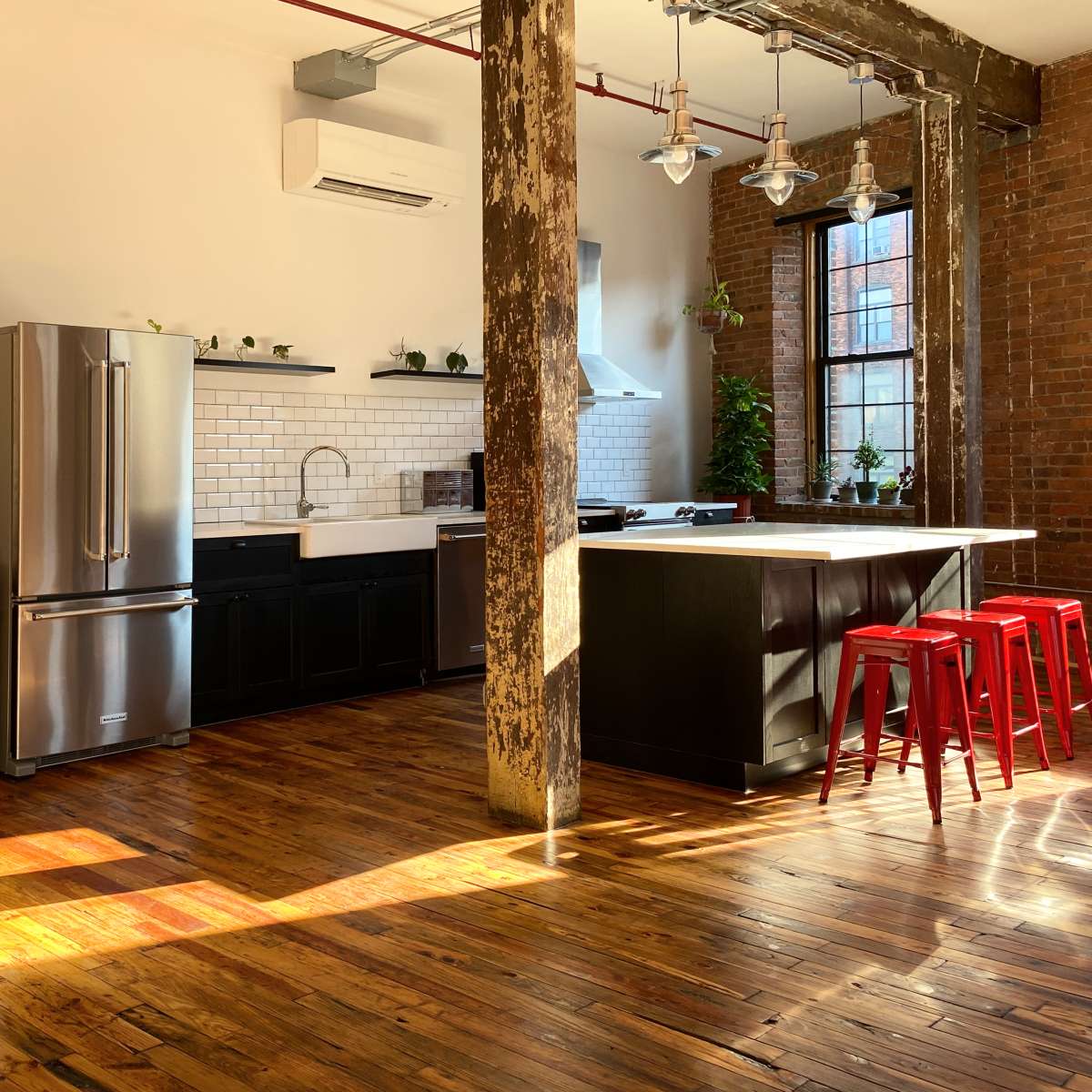 A modern kitchen with exposed brick walls, wooden flooring, stainless steel appliances, and red metal stools positioned at a kitchen island, alongside a large mirror reflecting part of the space.