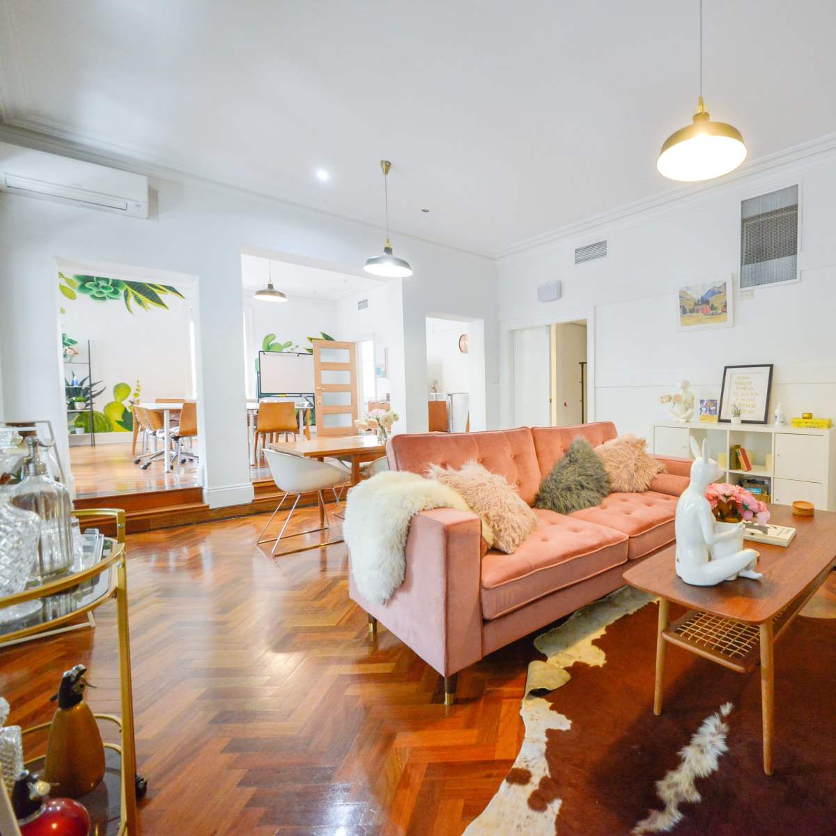 A living room with a pink sofa, wooden coffee table, and a decorative rug, featuring light-colored walls and a plant design in the background.